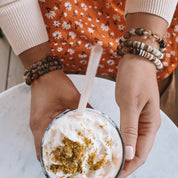 Person holding a drink with whipped cream and a straw, wearing bracelets, against a floral-patterned garment.