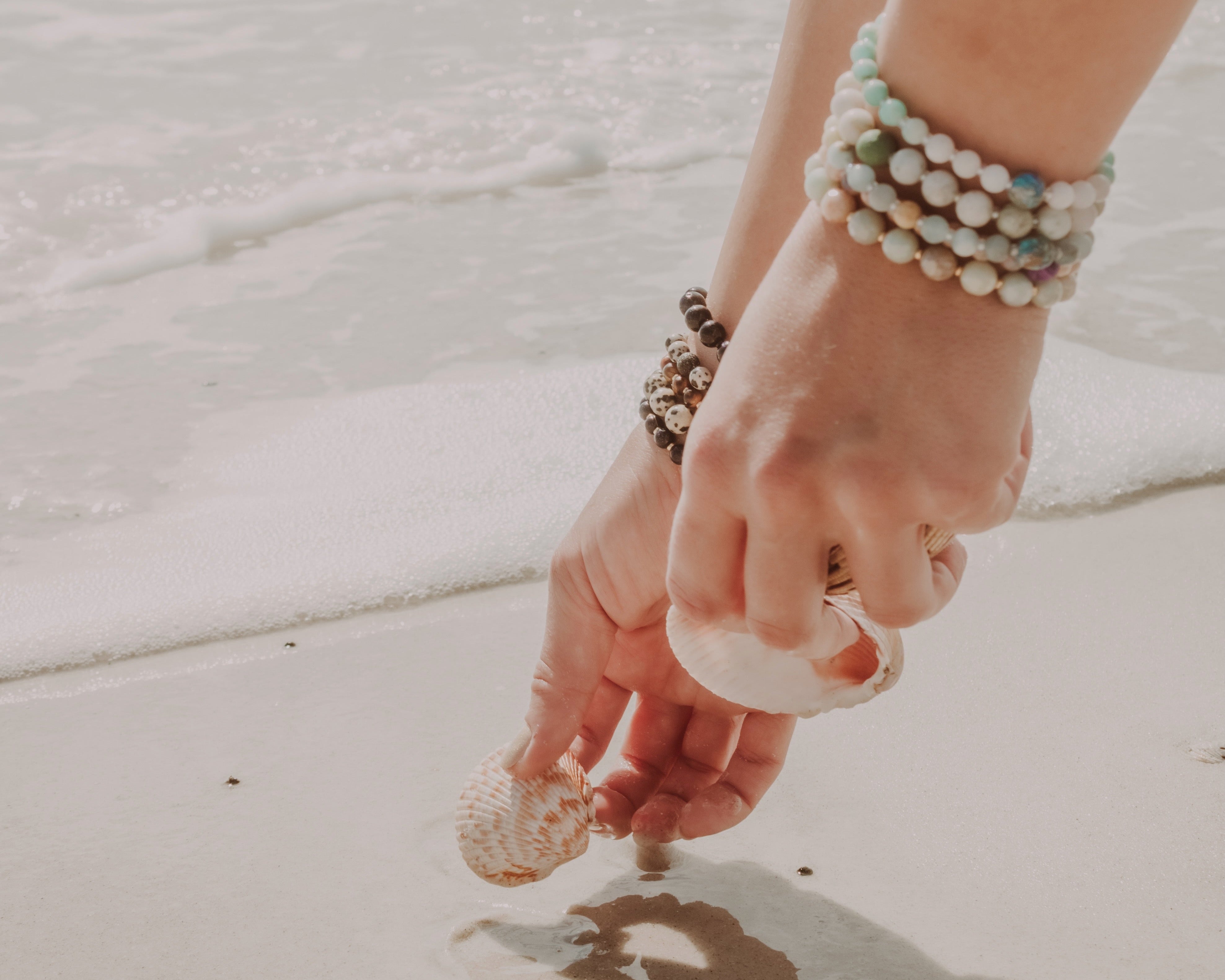 Person holding a shell on a beach with multiple bracelets on wrist